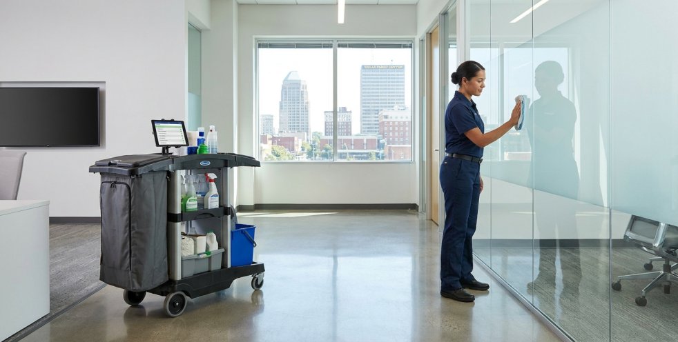 Commercial cleaner wiping a glass wall in a modern office with city skyline views and cleaning cart nearby